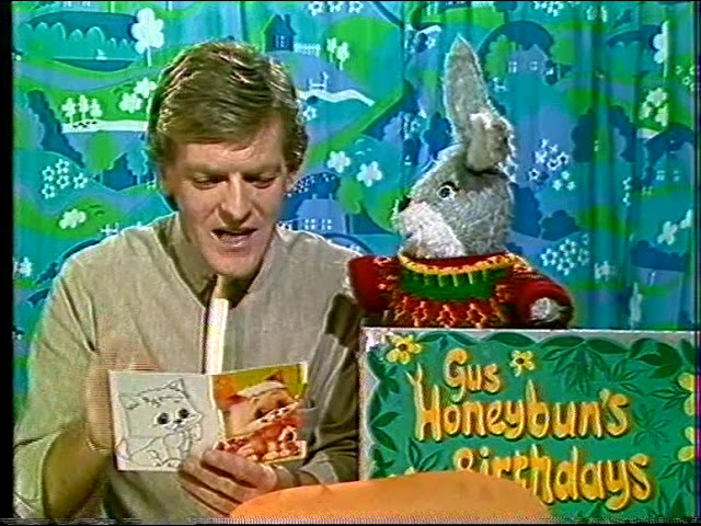 Ian Stirling and Gus Honeybun appearing on Westward Television. Ian Stirling is on the left of the picture, reading a birthday card with a kitten on the front. Gus is wearing a "Ted Tuckerman" fisherman’s jersey and looking at Ian. They are sat in front of a patterned blue, navy, green and white curtain. 